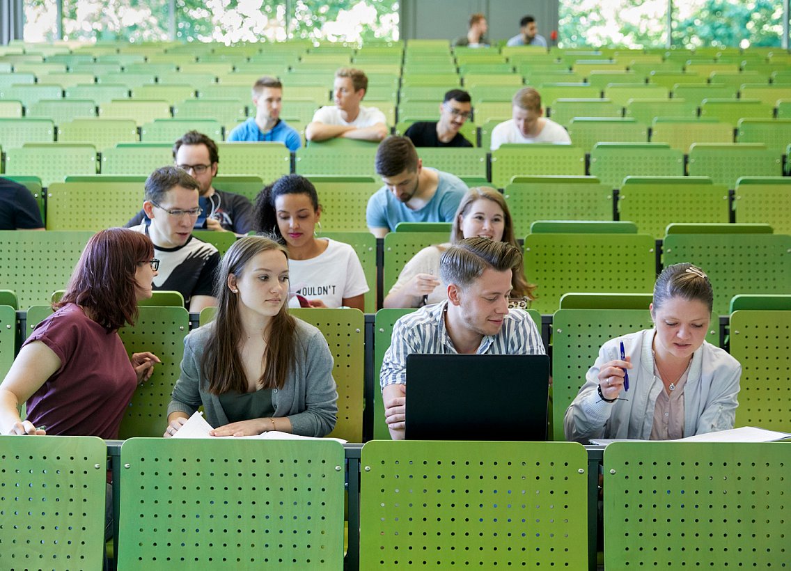 Studierende sitzen in einem Hörsaal der Uni Wuppertal, welcher grüne Stühle hat. 